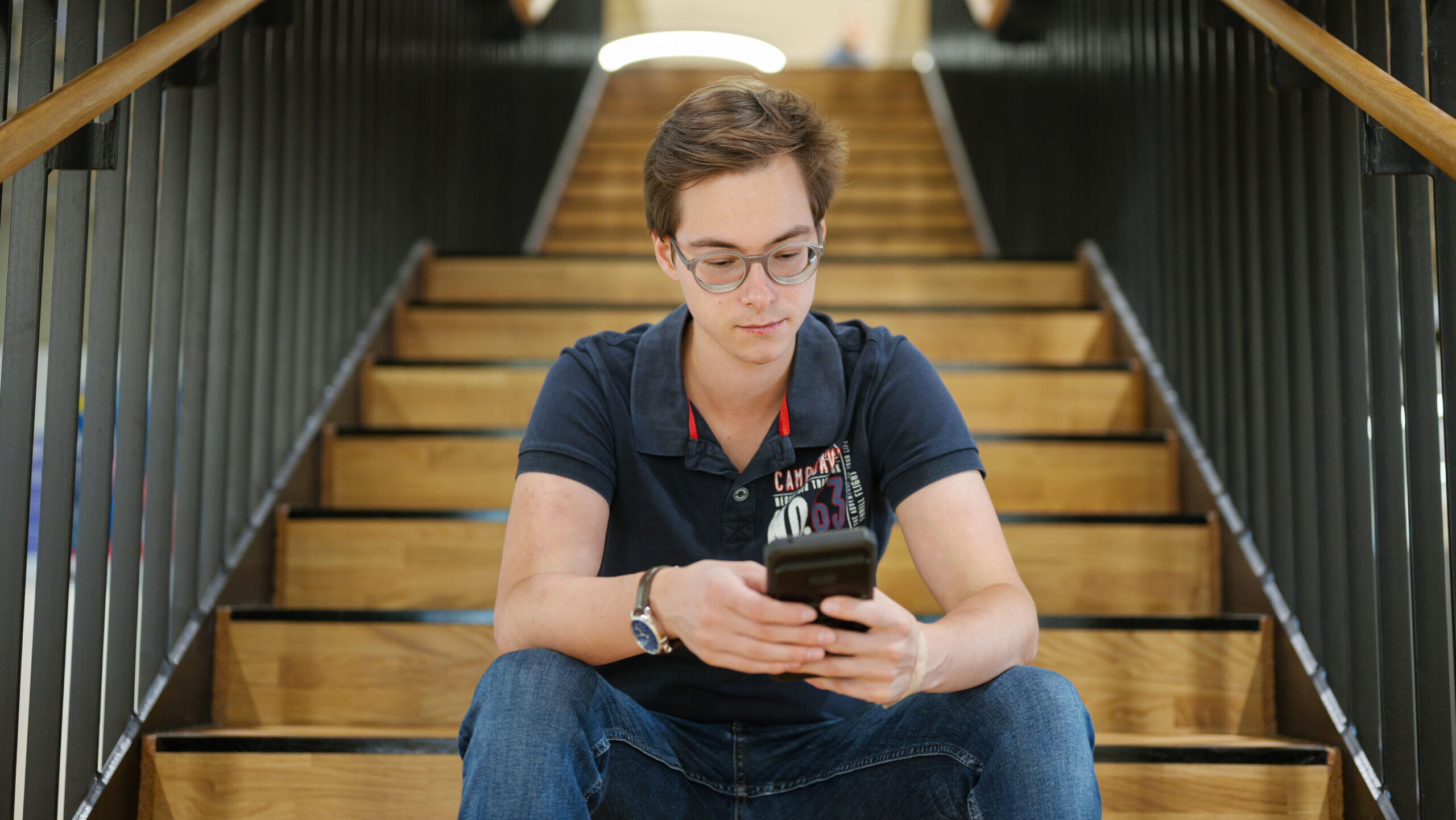 Ein junger Mann mit Brille und einem dunkelblauen Poloshirt sitzt auf einer Holztreppe und schaut auf sein Smartphone. Im Hintergrund sind weitere Stufen und ein Treppengeländer zu sehen.